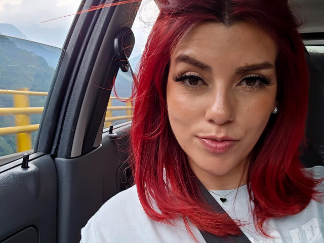 ZoeeMar with red hair, brown eyes, and a slight pout, wearing a white shirt, inside a car with a yellow railing outside, looking directly at the camera.