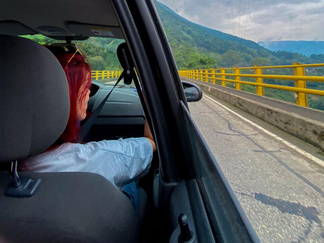 ZoeeMar with red hair, wearing a white shirt, sitting in a car, back to the camera, road and yellow guardrails in front, green hills and cloudy sky outside.