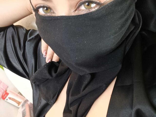 ZahraJalid has brown eyes, wearing a black mask covering her nose and mouth, black shirt, showing cleavage, with pink nail polish on her fingers.