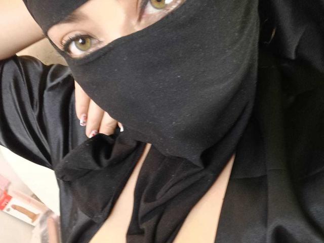 ZahraJalid with yellow eyes, wearing a black mask and robe, exposing cleavage, hand with pink nail polish resting on chest. Her fingers are slightly curled.