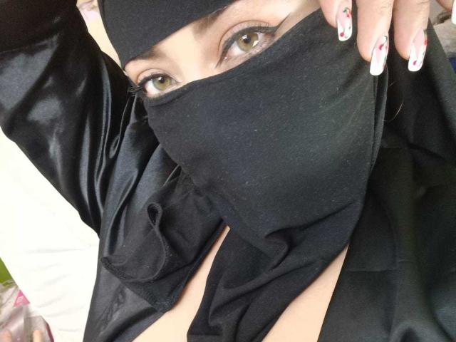ZahraJalid with a black mask covering her mouth and nose, wearing a black shirt, exposing her cleavage, and showing her green eyes with white and pink manicured nails.