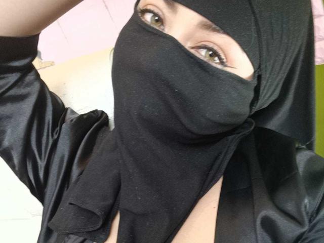 ZahraJalid with black mask covering her mouth and nose, black headscarf, and black satin shirt, exposing her brown eyes and part of her chest. Her hand's in the frame.