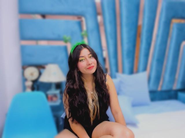 Genesiss with long dark hair, green eye makeup, black sleeveless top, and a smile, sitting on a blue chair, her bare legs and small tits visible.