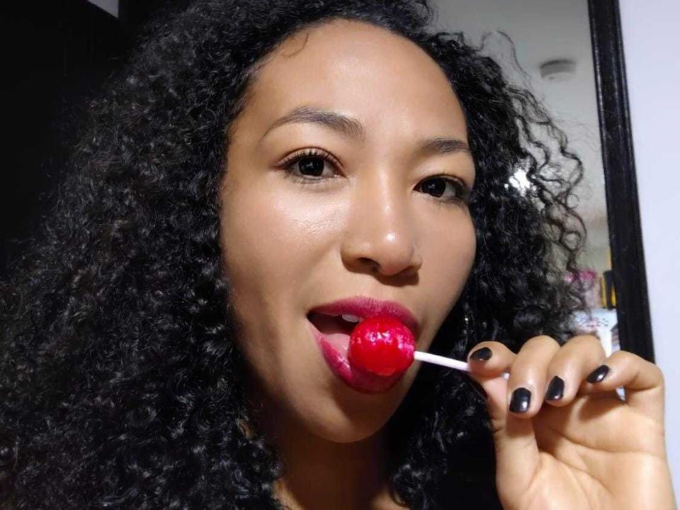 Yhulia with curly black hair, brown eyes, and pink lipstick, holding a red lollipop in her black-nailed hand, slightly open mouth, looking at the camera.