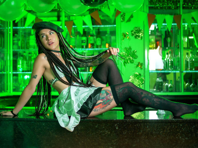 HellenK with long braids, black hat, and white skirt, showing off tattoos, black thigh-highs, and a sideboob, leaning on a shiny green-lit bar. Her ass is partially visible.