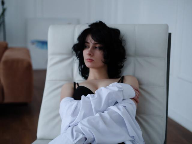 JulisSweet with wavy black hair, wearing a black bra and white shirt, arms crossed, sitting on a white leather chair, looking serious, boobs slightly visible.