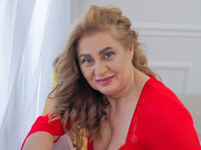 KarlaHartley, with wavy brown hair, wearing a red lace top, smiling slightly, showing her tits, has pink lipstick, and is looking directly at the camera.