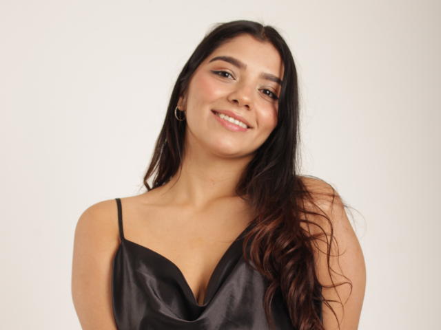Issanassar with long brown hair, smiling, wearing a black satin dress with thin straps, showing her small tits, against a plain white background.