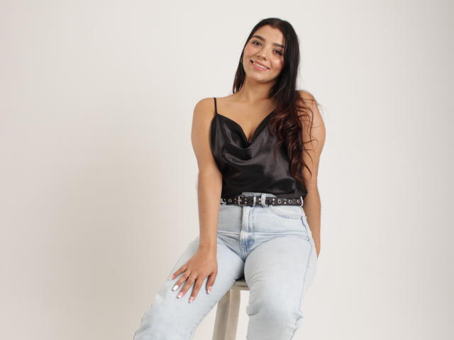 issanassar with long dark hair, smiling, wearing black satin top, light blue jeans, and black belt, sitting on stool, hands on legs, looking at camera, cute, sexy.
