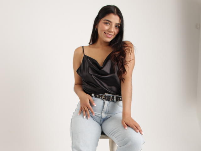 issanassar sitting on a stool, wearing a black satin top with thin straps, light blue ripped jeans, and a black belt with silver studs, smiling at the camera.