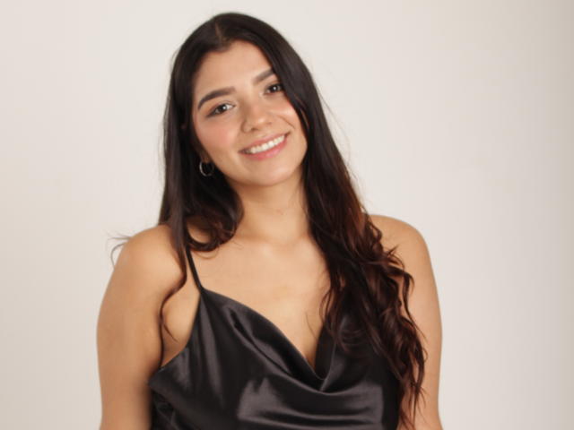 Issanassar with long dark brown hair, smiling, wearing a black satin dress with a deep V-neck, small gold hoop earrings, light skin, and brown eyes.