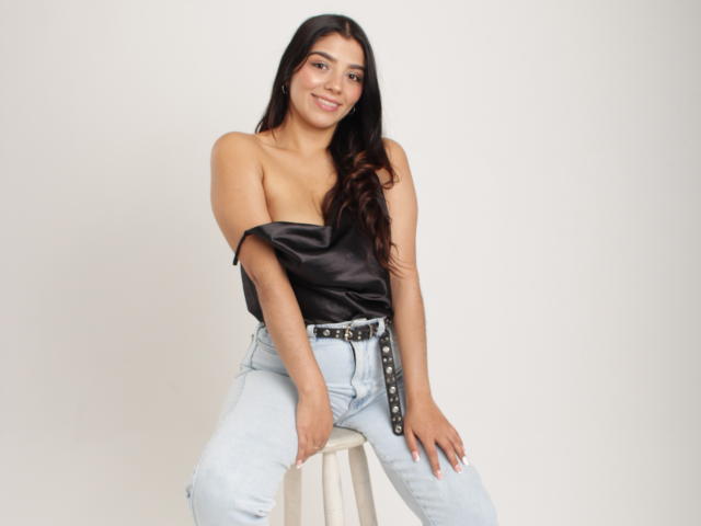 Issanassar with long dark hair, smiling, wearing a black off-shoulder top, light blue jeans, and a studded belt, sitting on a stool. Boobs visible.