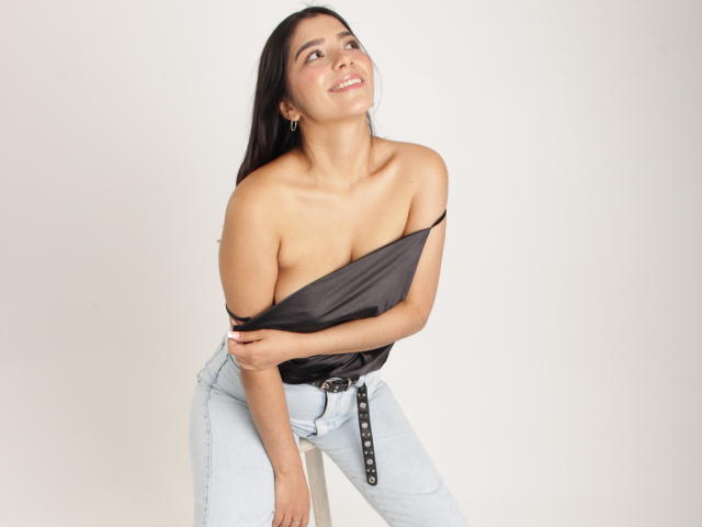 Issanassar with dark hair, smiling, has her black top pulled down to expose her tits, wearing light blue jeans with a black belt, against a white background.