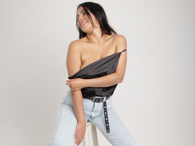 Issanassar with long black hair, smiling, wearing a black off-shoulder top, light blue high-waisted jeans, and a black belt, sitting on a stool, showing her tits.