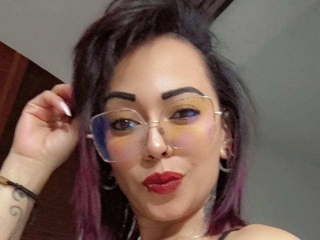 IsaHot69 with glasses, purple-dyed hair, and red lipstick, touching her hair, wearing a nose ring, and hoop earrings, showing off her tattooed arm.