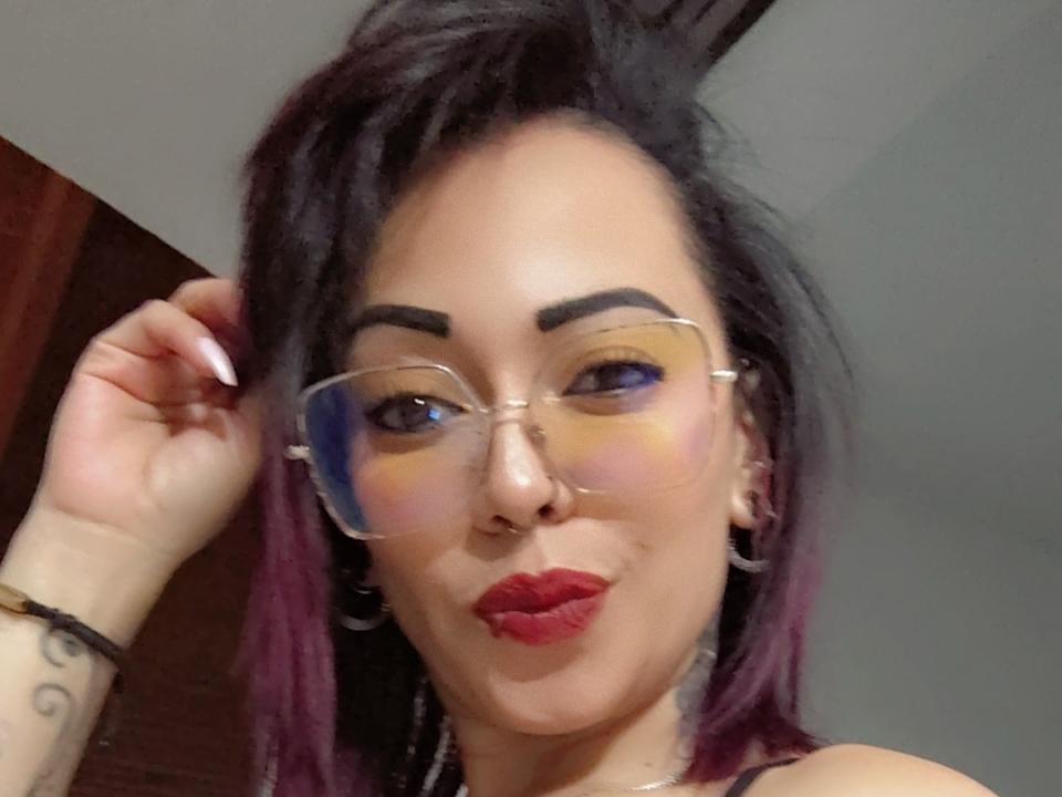 IsaHot69 with glasses, purple-dyed hair, and red lipstick, touching her hair, wearing a nose ring, and hoop earrings, showing off her tattooed arm.