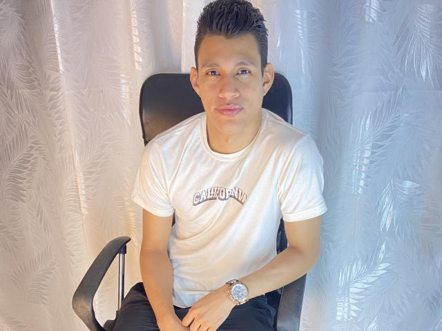 TheNewMan, ein asiatischer Mann mit kurzen, schwarzen Haaren, der ein weißes T-Shirt mit der Aufschrift "California", schwarze Hosen und eine Uhr trägt, sitzt auf einem schwarzen Stuhl und zieht die rosa Lippen zusammen..