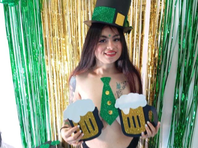 MiaCollivens with long brown hair, green tie, and black top hat, holding glittery beer mugs, exposing her bare tits, wearing a green shamrock on her chest. Stupid grin.