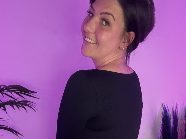 NatiLoveYou, souriant(e) avec les dents visibles, chemise noire à manches longues, cheveux bruns foncés attachés en chignon, arrière-plan illuminé de violet, plante sur la gauche, petits boucles d'oreilles..