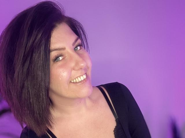 NatiLoveYou has short dark brown hair, blue eyes, and a slight smile showing teeth. She's wearing a black top with a visible black bra strap. Purple background.