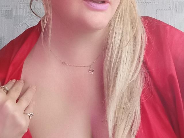 KelliRedhaer's face and chest, showing pink lipstick, blonde ponytail, red satin robe, gold necklace with small pendant, and hand with rings, gently touching her chest.
