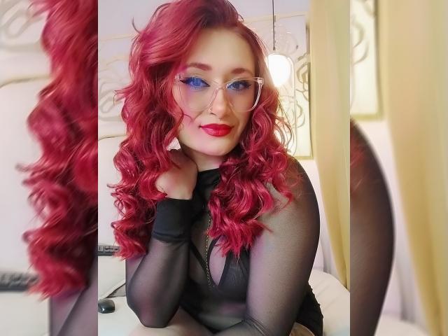PrincesaLilith with red curly hair, wearing transparent black long-sleeve top, clear glasses, and red lipstick, sitting with hands near face, smiling at camera. Boobs visible through top.