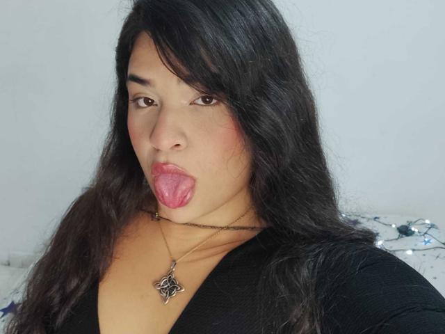 AntonelaPark with long black hair, brown eyes, and her tongue out, wearing a black top and a necklace with a star pendant, showing a bit of her chest.
