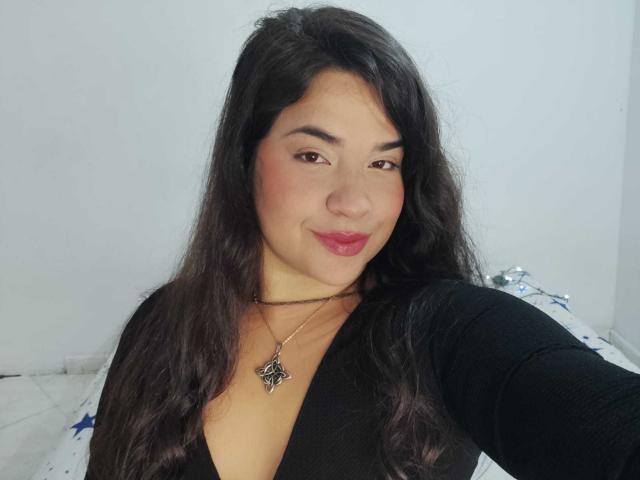 AntonelaPark with long black hair, wearing a black V-neck, has a necklace with a pentagram, smiling with red lipstick, her tits partially visible, white background.