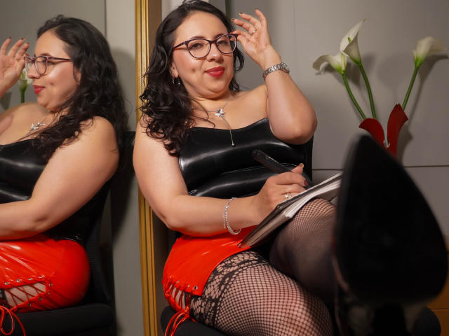 Lisa Teylor mit lockigem, schwarzem Haar, Brille, rotem Lippenstift, einem schwarzen, trägerlosen Lederoberteil, roten Schnürhosen, Fischernetzstrümpfen, schwarzen High Heels, hält ein Buch, sitzt, Spiegelbild..