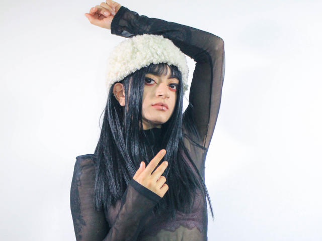Amelie Sato, con capelli neri, un cappello bianco e peloso, una maglietta nera trasparente, un braccio sollevato, il dito medio alzato, pelle pallida e trucco rosso, con un'espressione seria. Il seno è visibile attraverso la maglietta..