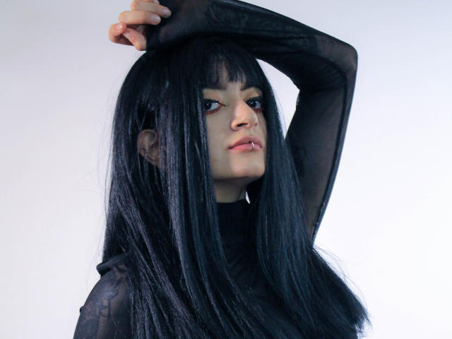 AmelieSato with long black hair, wearing a sheer black top, has her arm raised, showing off her tits and a slightly annoyed expression, against a white background.