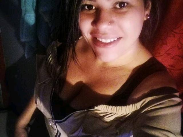 AlmaWhite69 with dark hair, nose ring, and earrings, smiling at camera, wearing unzipped gray top, showing black bra and cleavage, standing in dimly lit room.