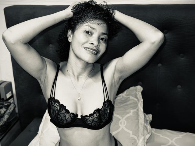 Foto em preto e branco de Caelina com cabelo cacheado, sorrindo, com os braços levantados, vestindo um sutiã de renda, seios pequenos, mãos no cabelo, colar, sentada na cama com travesseiros..