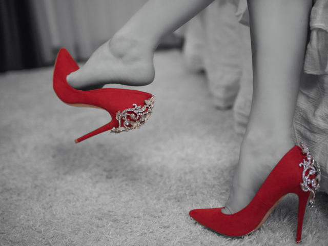 ReinaStarx's legs in red high heels with silver embellishments, one foot lifting the other, standing on a gray carpet, showing off her sexy, stylish, and elegant footwear.