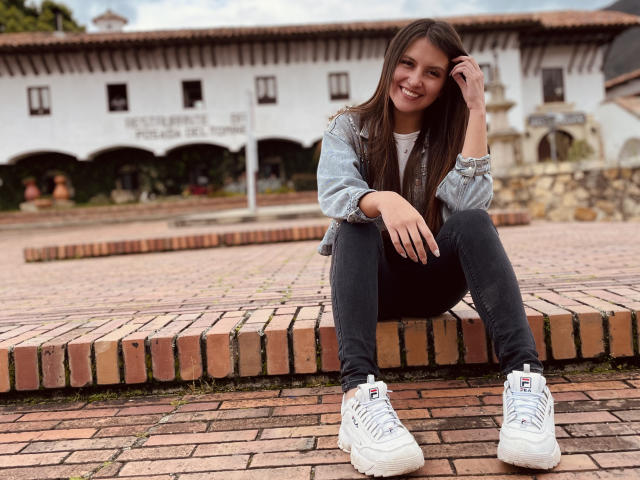 AmelieMuller, sitting on brick ledge, smiling, hand in brown hair, wearing gray jacket, black pants, white sneakers, long brown hair, cute, attractive, outdoor, overcast, casual, relaxed.