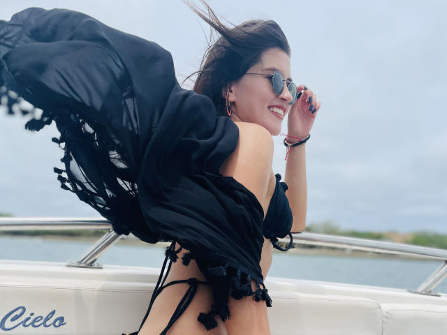AmelieMuller with black sheer shawl, black bikini, and sunglasses, smiling, showing her tits, ass, and small black thong, standing on a boat, wind in her hair.