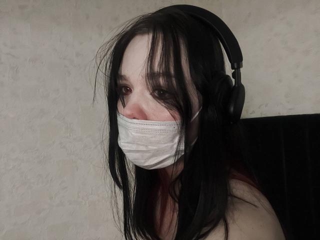 ViellaRosewood with black hair, wearing a white mask, headphones, and nothing on her shoulders, looking slightly to the side, against a plain wall.