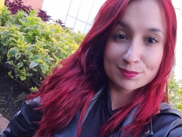 Bellanid has red hair, a cute face, and pink lipstick. She's wearing a black leather jacket, and her ass is not visible. She's smiling with a slight nose bump.