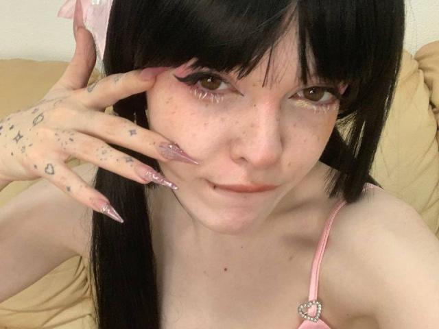 Azusa Nakano, con capelli neri, eyeliner rosa e unghie lunghe rosa, mostra dei tatuaggi sulla mano, indossa lingerie rosa con un fiocco e ha un leggero sorriso, seduta su un divano beige..