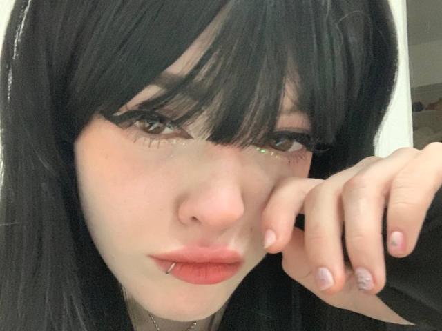 AzusaNakano has black hair, piercings on her lower lip, and glittery eyeliner. She's resting her chin on her hand with pink nail polish. Her eyes are half-closed.