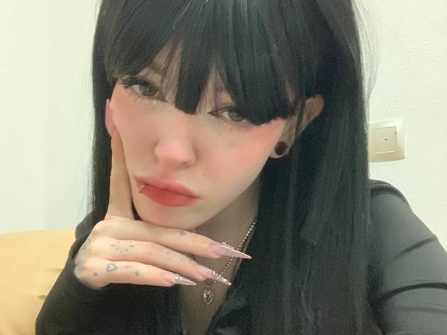Azusa Nakano hat schwarzes Haar, eine Ponyfrisur und einen Nasenring. Sie trägt ein schwarzes Hemd, hat lange, rosafarbene Nägel und berührt ihre Wange mit tätowierten Fingern..
