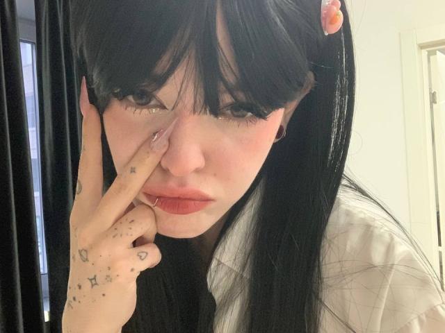 Azusa Nakano, avec ses cheveux noirs, sa frange et son anneau de nez rose, touchant ses lèvres avec un doigt, montrant des tatouages en forme d'étoiles sur sa main, portant une chemise blanche, regardant la caméra..