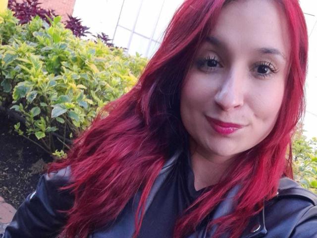 Bellanid with red hair, black leather jacket, and pink lipstick, smiling at the camera, showing her face and upper body, with green plants in the background.