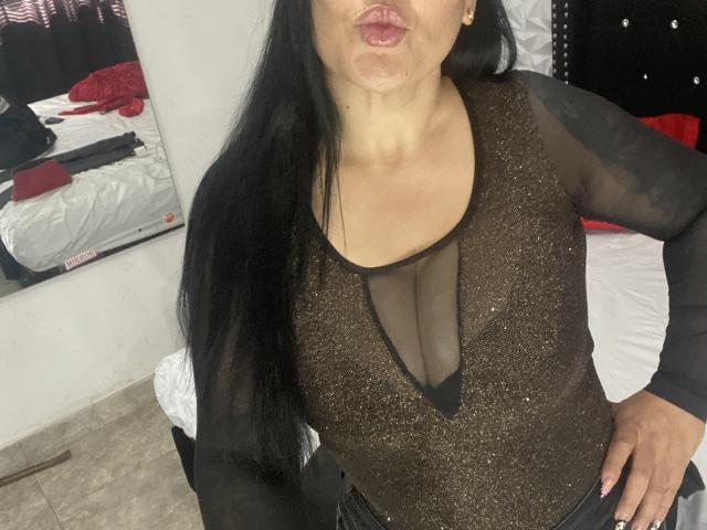 KittyHotLady69 mit langen, schwarzen Haaren, trägt ein glitzerndes, braunes Oberteil mit tiefem Ausschnitt, das ihre mittelgroßen Brüste, vollen Lippen und eine Hand, die auf der Hüfte liegt, zeigt..