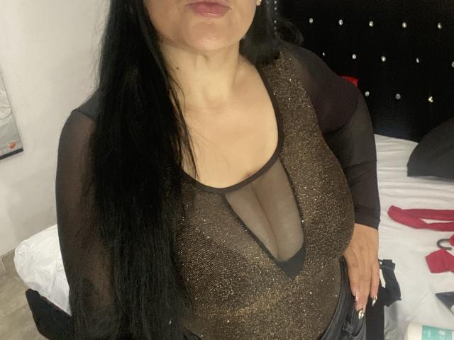 KittyHotLady69 con lunghi capelli neri, indossa una maglia trasparente marrone con un profondo scollo, che mette in mostra il suo seno generoso e una parte della sua cintura nera..