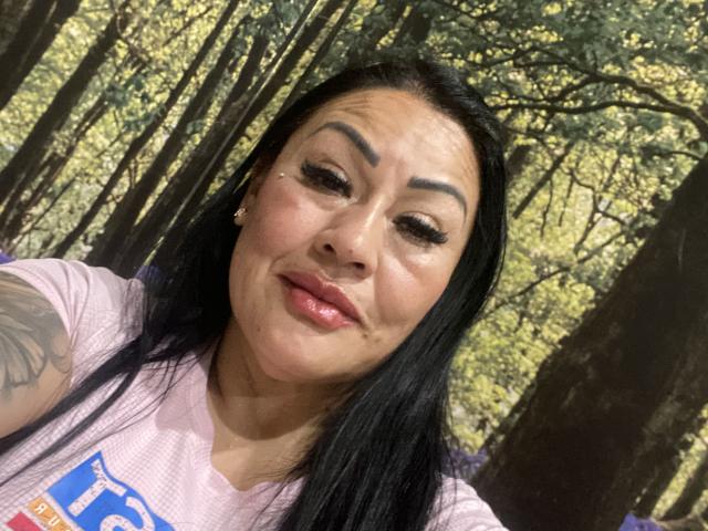 KittyHotLady69 with black hair, thick eyebrows, and a tattoo on her right arm, wearing a pink shirt, has shiny pink lips and a small beauty mark on her right cheek.