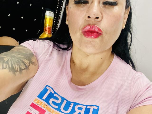 KittyHotLady69 con rossetto rosso lucido, camicia rosa gonfia, tatuaggio nero sul braccio destro, lattina di birra sullo sfondo, che fa una smorfia, tenendo il telefono per un selfie..
