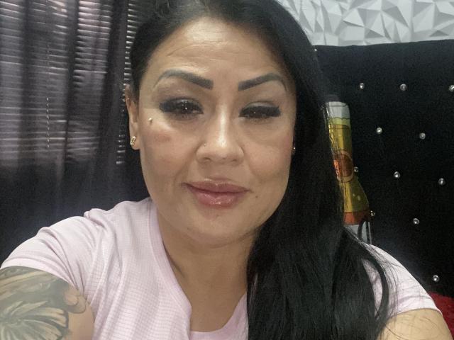 KittyHotLady69 a les cheveux foncés, des sourcils épais et un bras tatoué. Elle porte une chemise rose, a un anneau au nez et un léger sourire, avec une bouteille en arrière-plan..