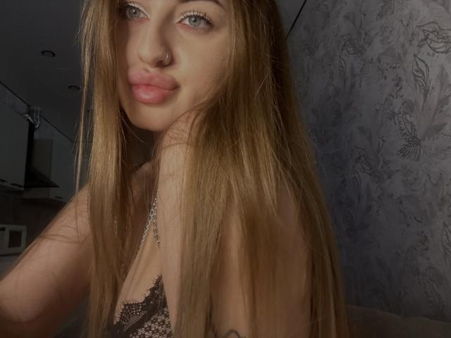 BabeCandy69 with long brown hair, pouty pink lips, nose ring, and lace top, showing her small tits in a low-lit room. Soft gray wallpaper in the background.