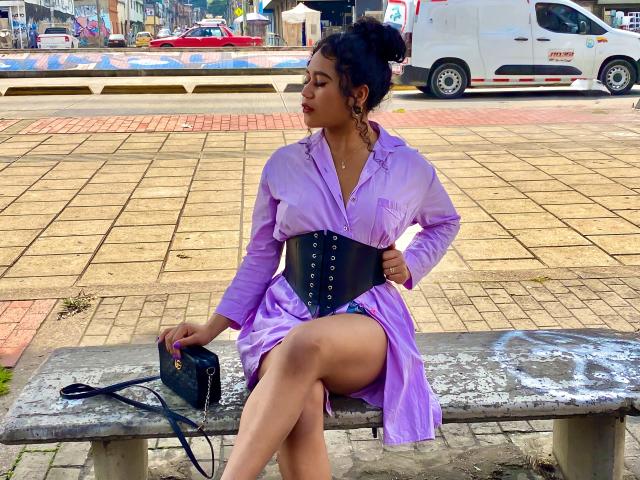 SophieSonyas with purple dress, black corset, and black purse, sitting on a bench, legs crossed, showing her ass, with her hair in a bun, in an urban setting.